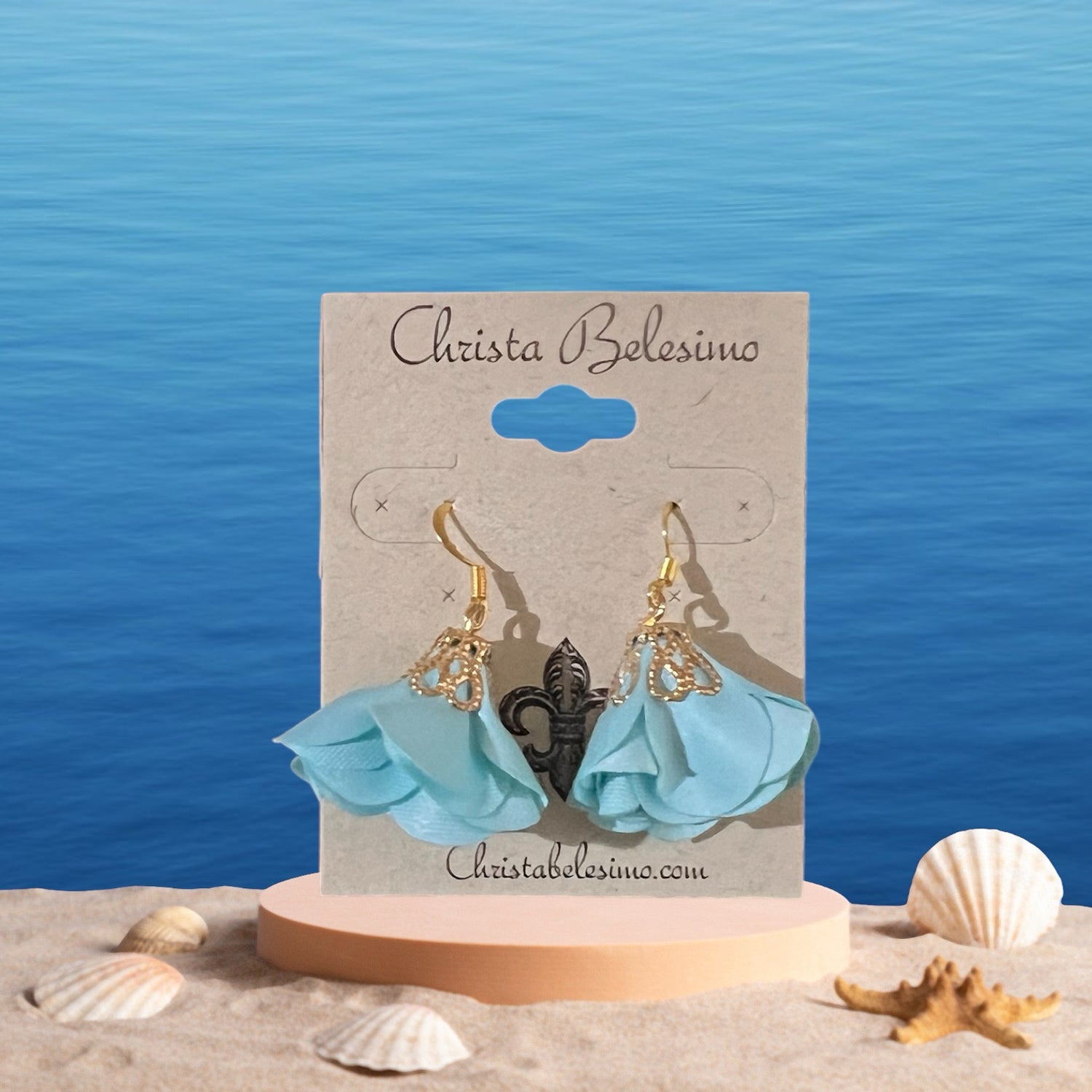 A pair of gold and blue satin flower earrings on a display card, with the brand name 'Christa Belisio'. The card is placed on a beach-like setting with shells and a blue background.