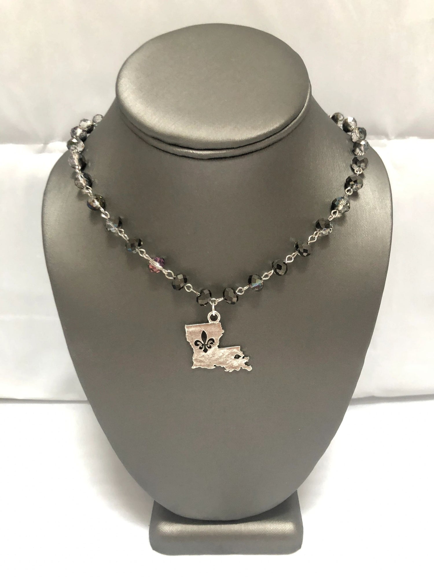 A silver-colored necklace with smoky grey beads and a fleur-de-lis pendant representing the state of Louisiana, displayed on a mannequin bust.