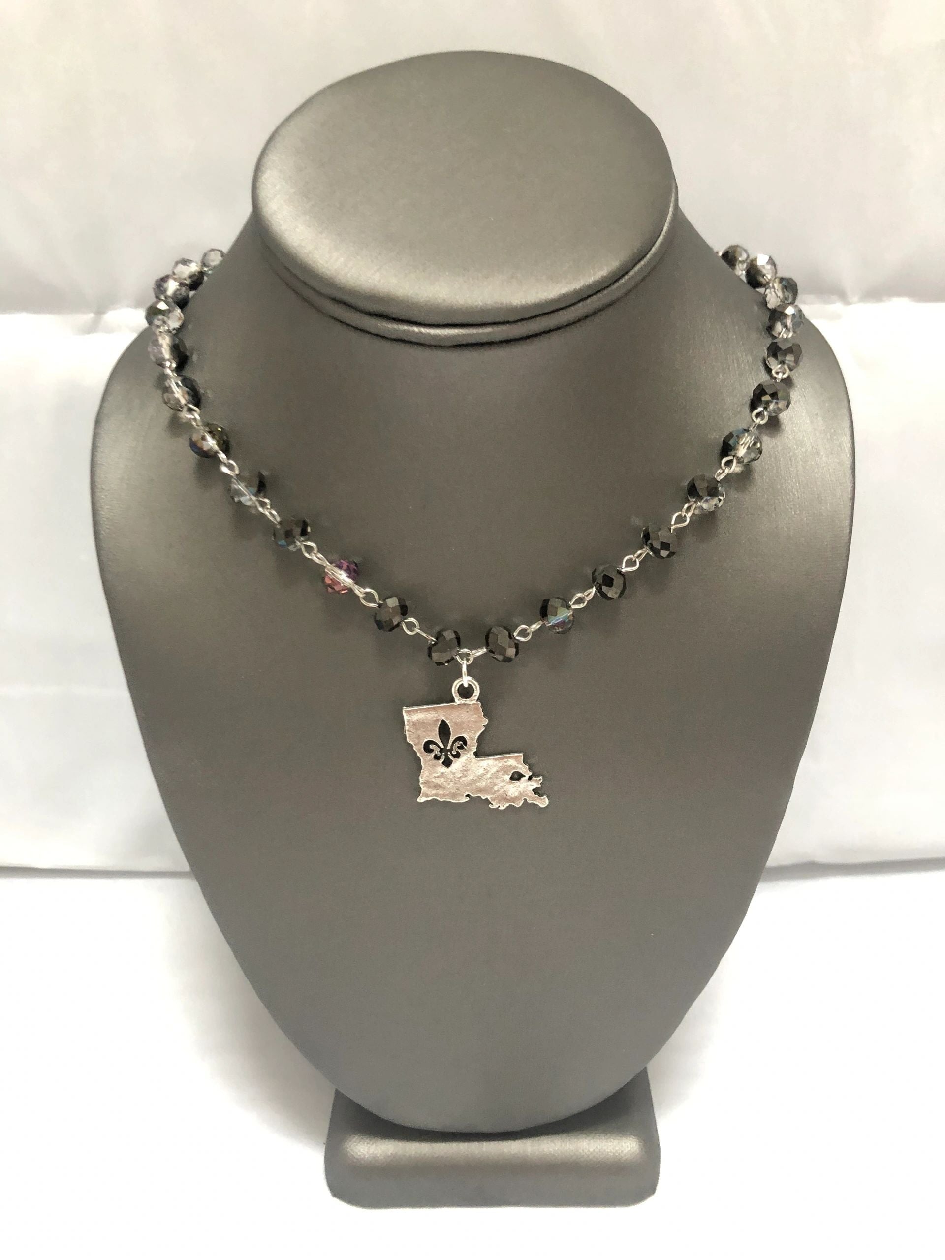 A silver-colored necklace with smoky grey beads and a fleur-de-lis pendant representing the state of Louisiana, displayed on a mannequin bust.