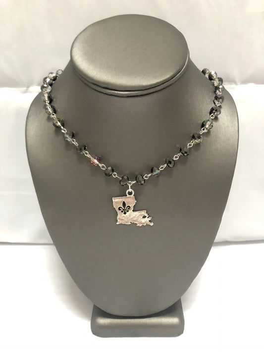 A silver-colored necklace with smoky grey beads and a fleur-de-lis pendant representing the state of Louisiana, displayed on a mannequin bust.