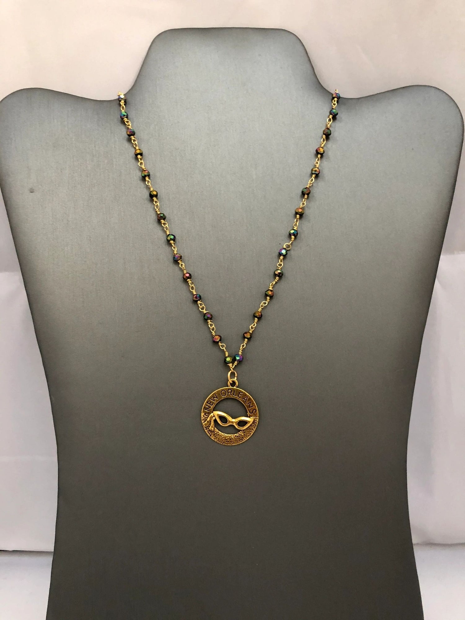 A beaded necklace with a gold-tone pendant featuring a Mardi Gras mask design, displayed on a mannequin.
