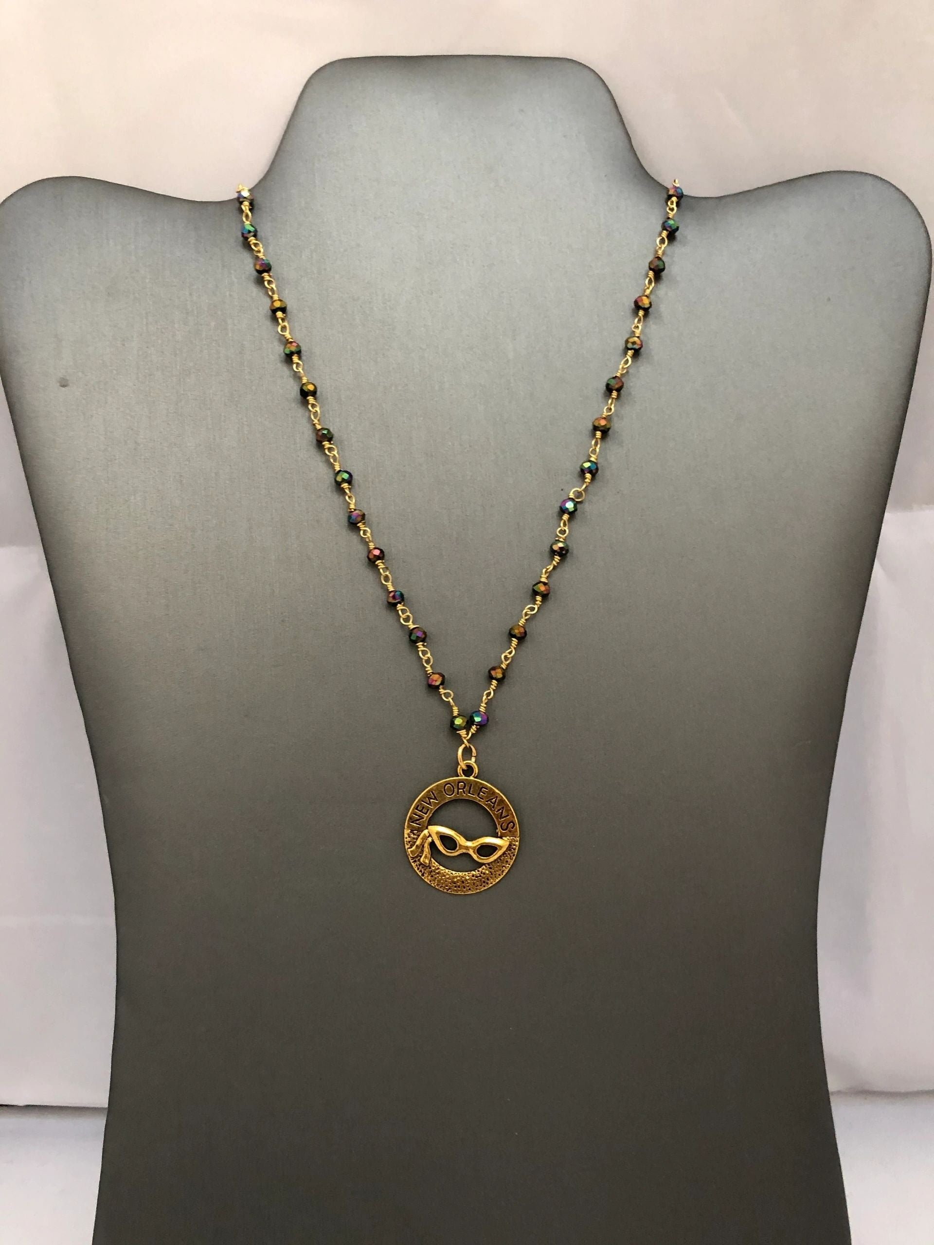 A beaded necklace with a gold-tone pendant featuring a Mardi Gras mask design, displayed on a mannequin.