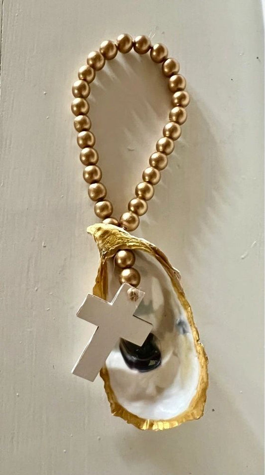 A gold-trimmed oyster shell with a white wooden cross and gold wood beads against a textured background.