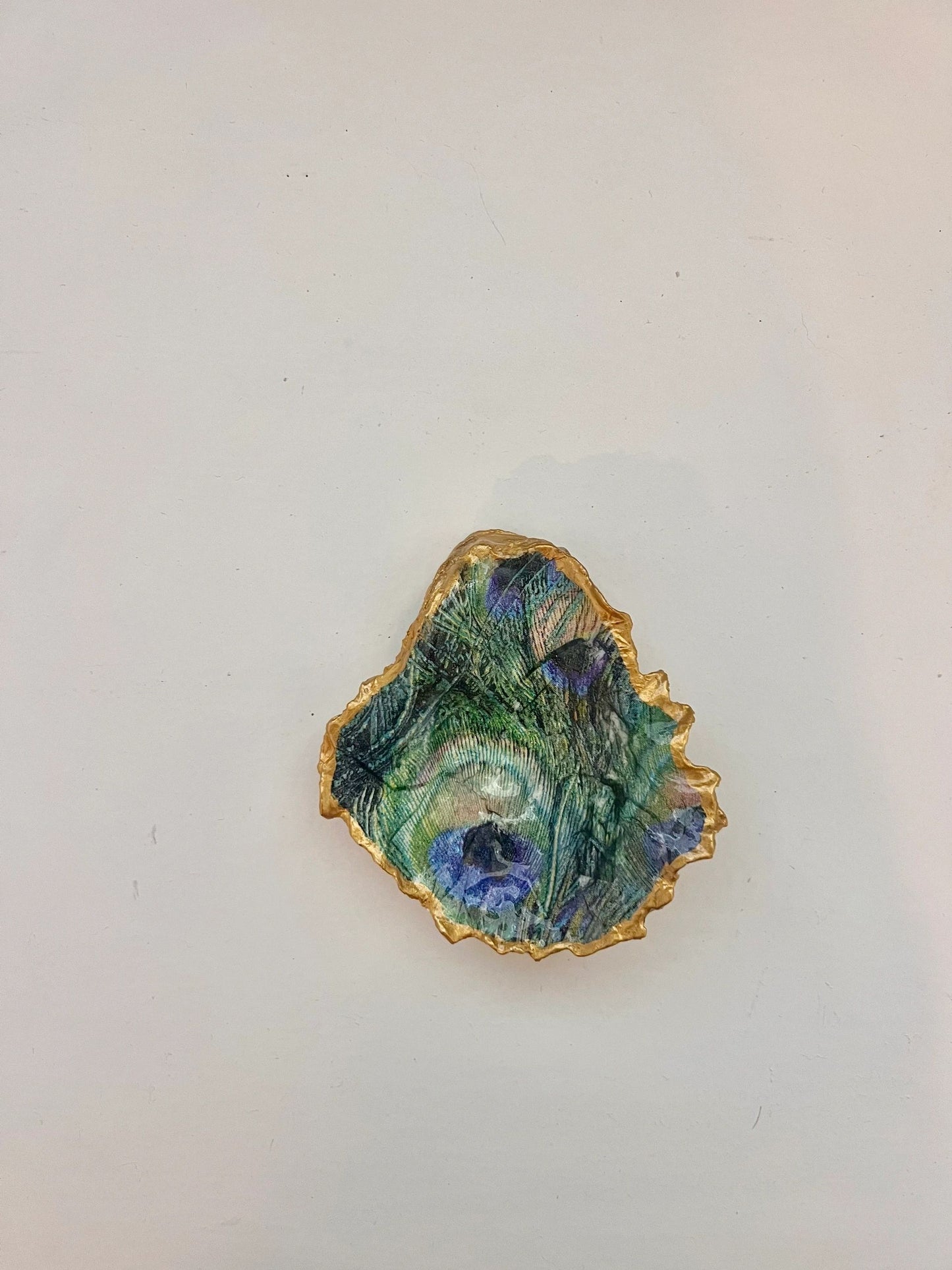 A gold-trimmed oyster shell trinket holder with a peacock feather pattern on the bottom right.