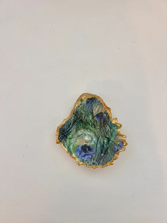A gold-trimmed oyster shell trinket holder with a peacock feather pattern on the bottom right.