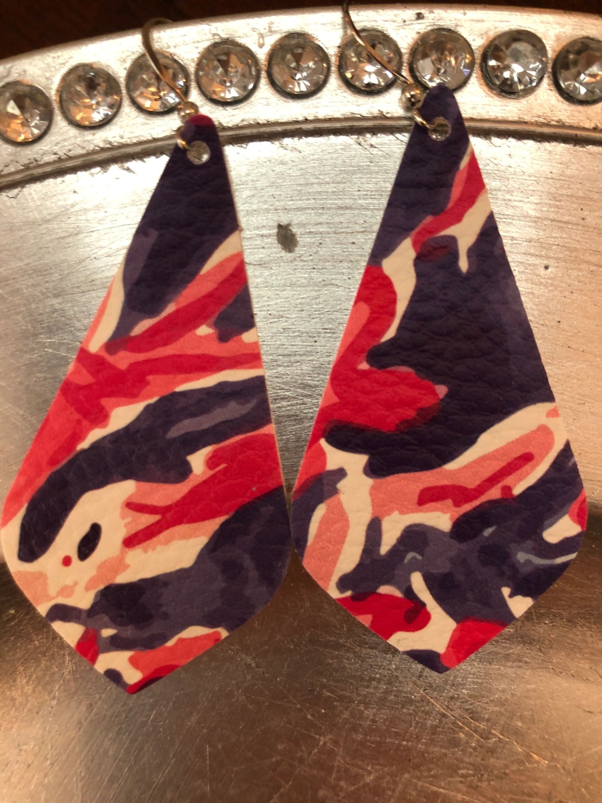 A pair of pink, blue, and purple leather earrings displayed on a reflective surface.