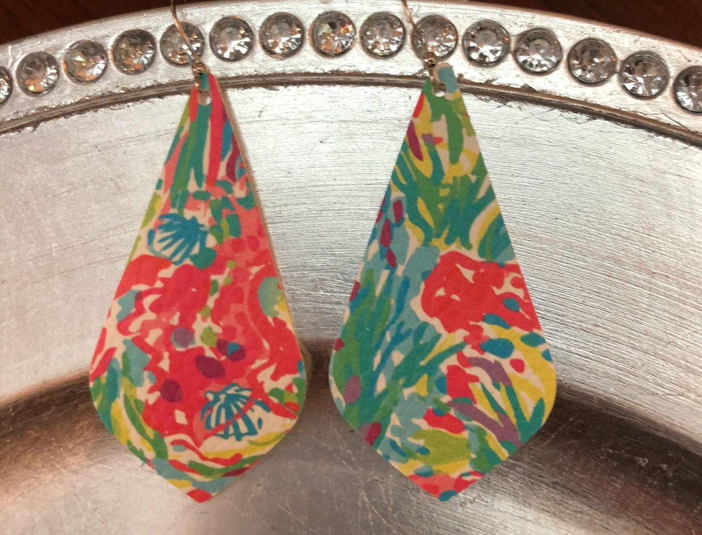 A pair of pink and turquoise floral print leather earrings displayed on a metal surface.