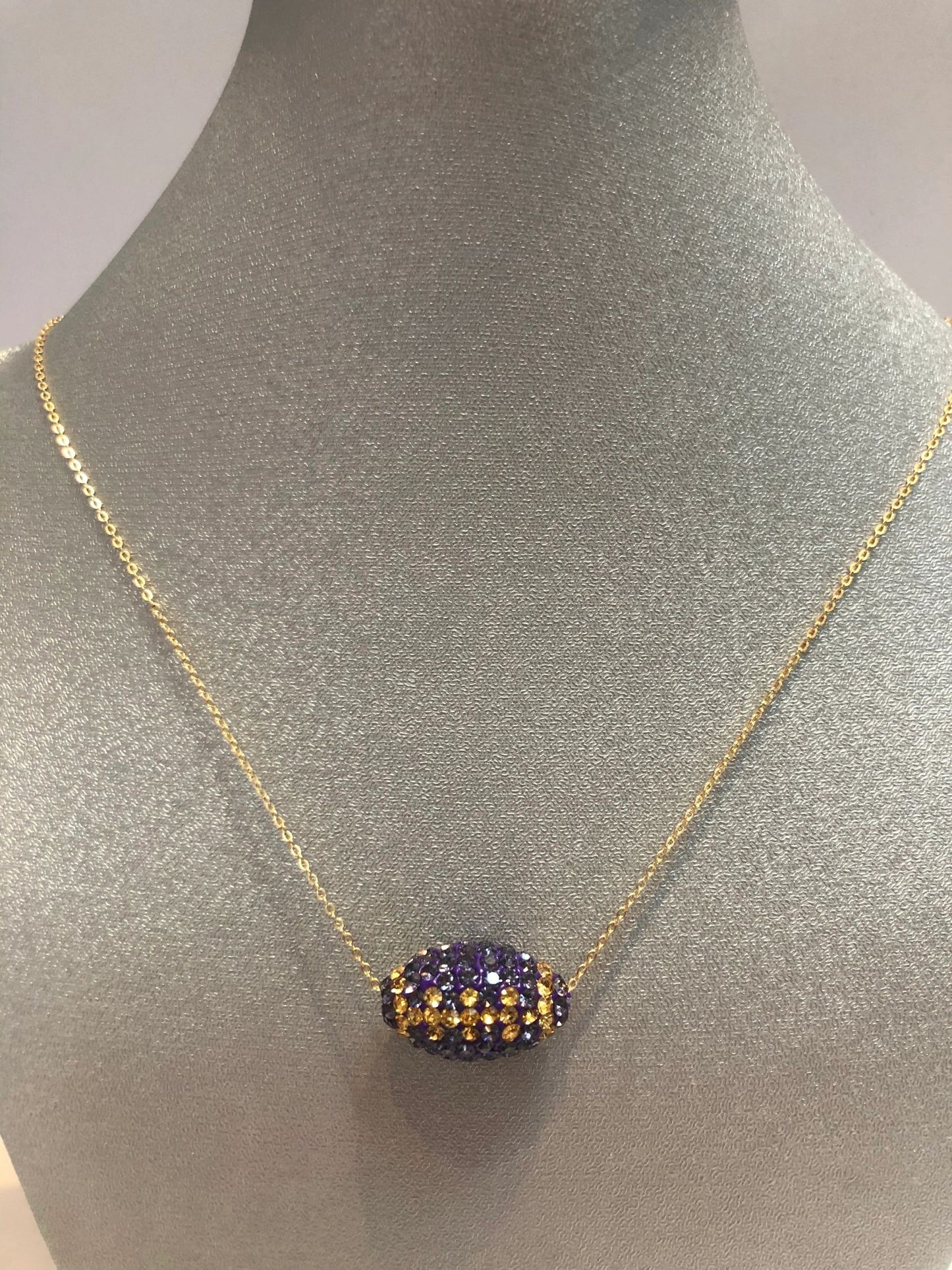 A gold-colored necklace with a football-shaped pendant featuring purple and gold crystals displayed on a mannequin bust.