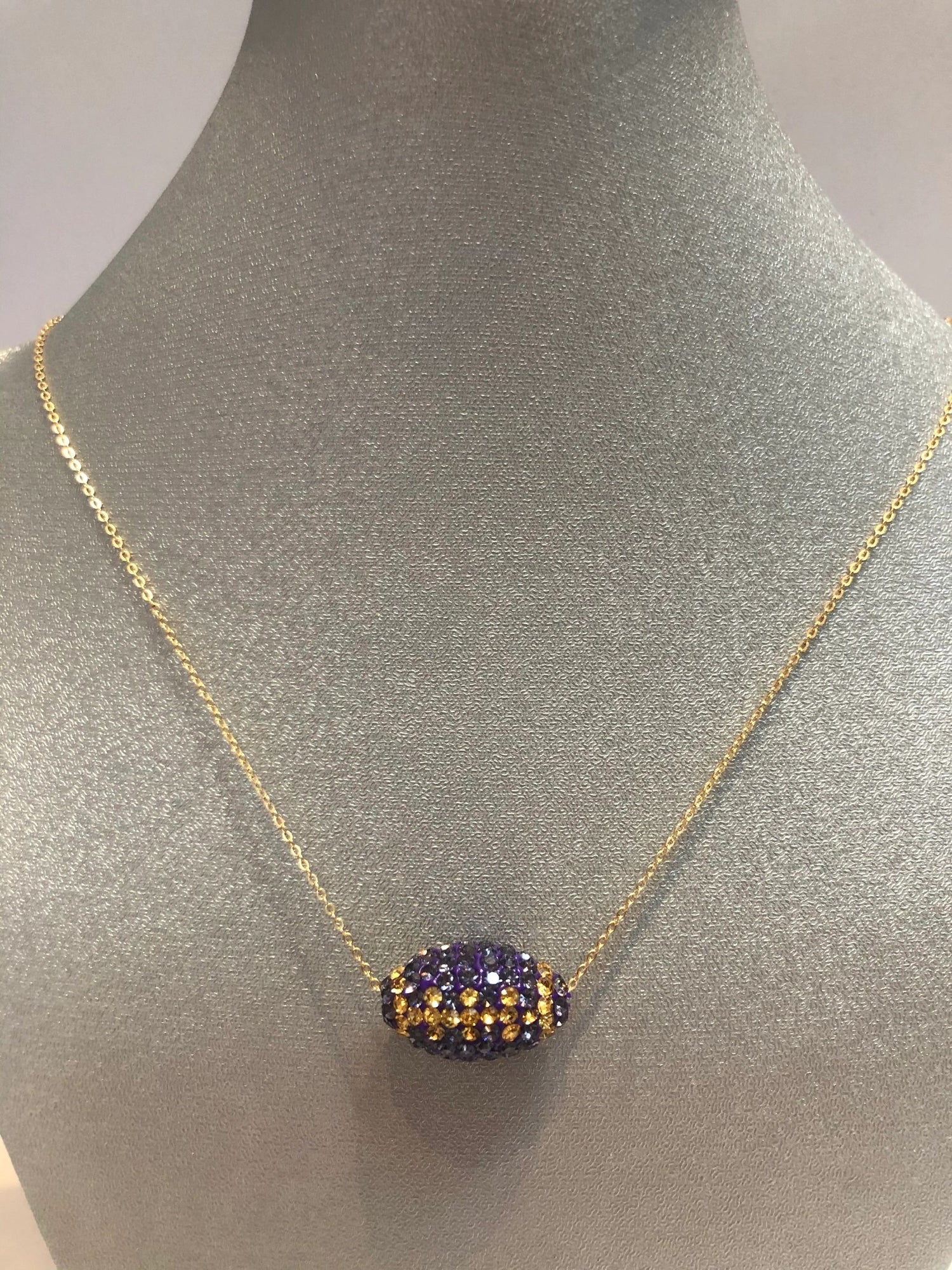 A gold-colored necklace with a football-shaped pendant featuring purple and gold crystals displayed on a mannequin bust.