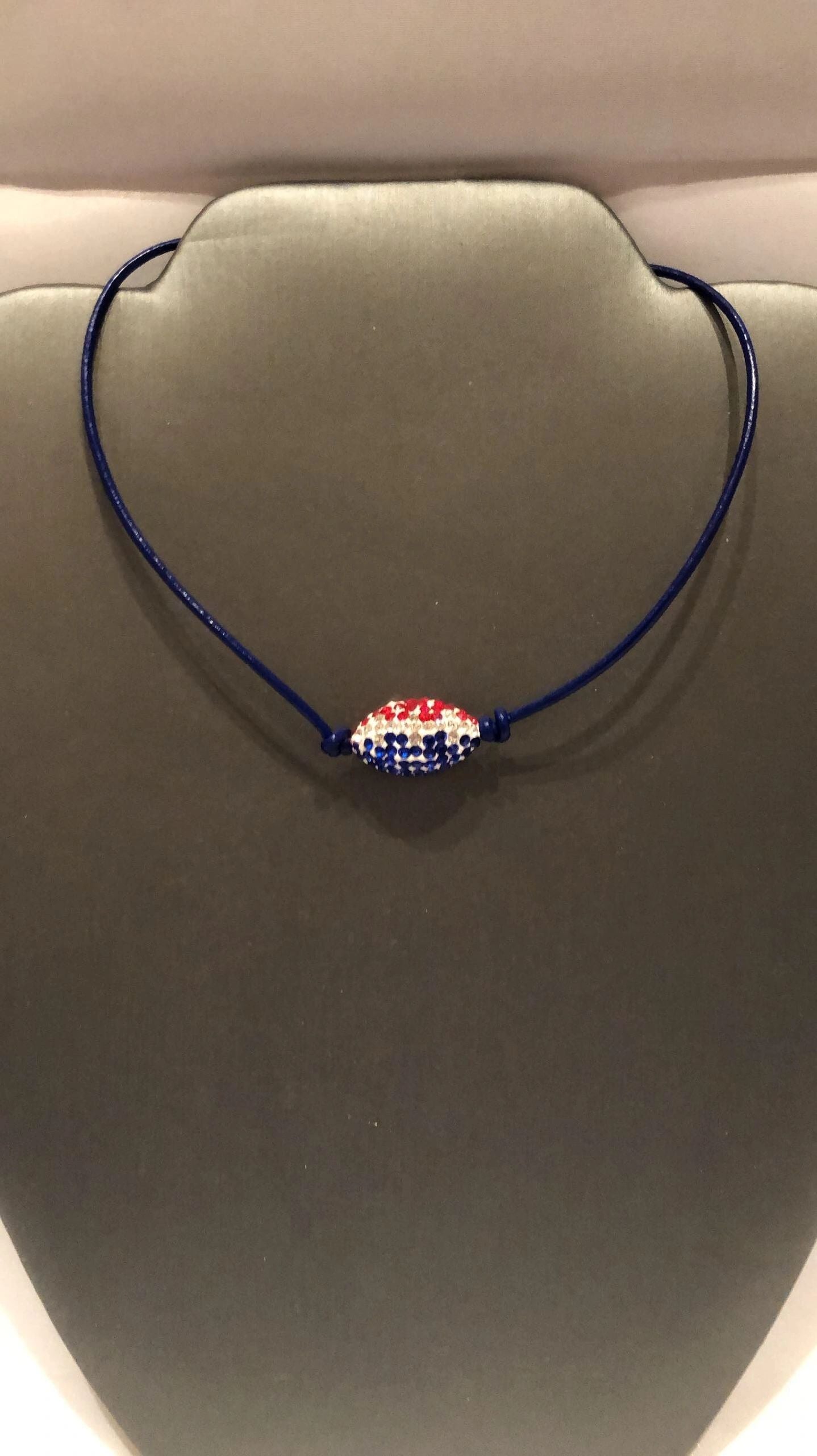 A blue leather cord necklace with a silver lobster clasp featuring a pave' football pendant with Patriot's or Houston Texan colors.