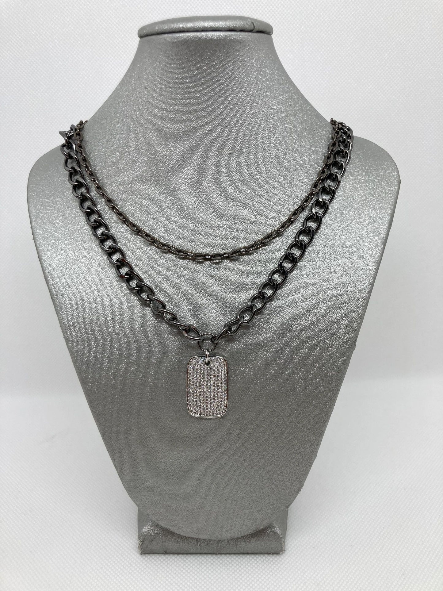 A silver pave dog tag pendant on a cuban link style double gunmetal chain, displayed on a mannequin bust.