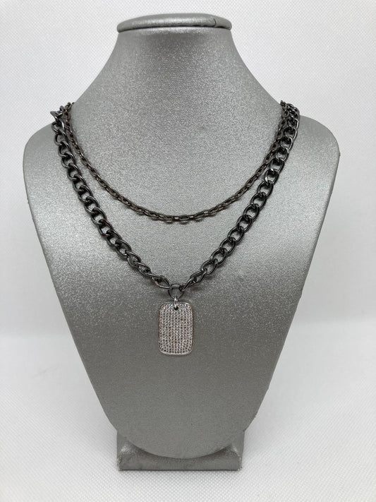 A silver pave dog tag pendant on a cuban link style double gunmetal chain, displayed on a mannequin bust.
