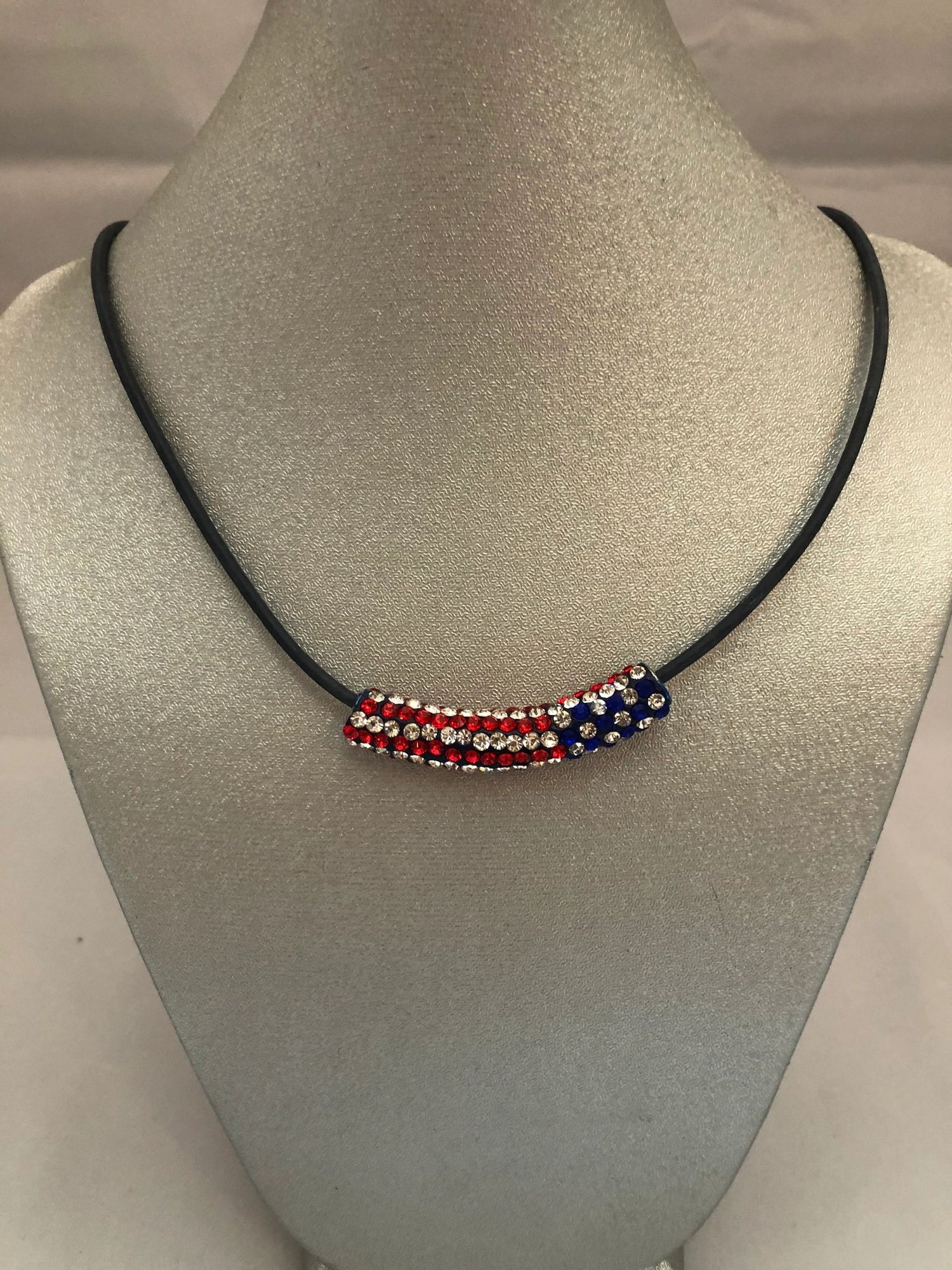 A necklace with a black leather cord and a pave’ crystal bar featuring a flag design in red, white, and blue colors.