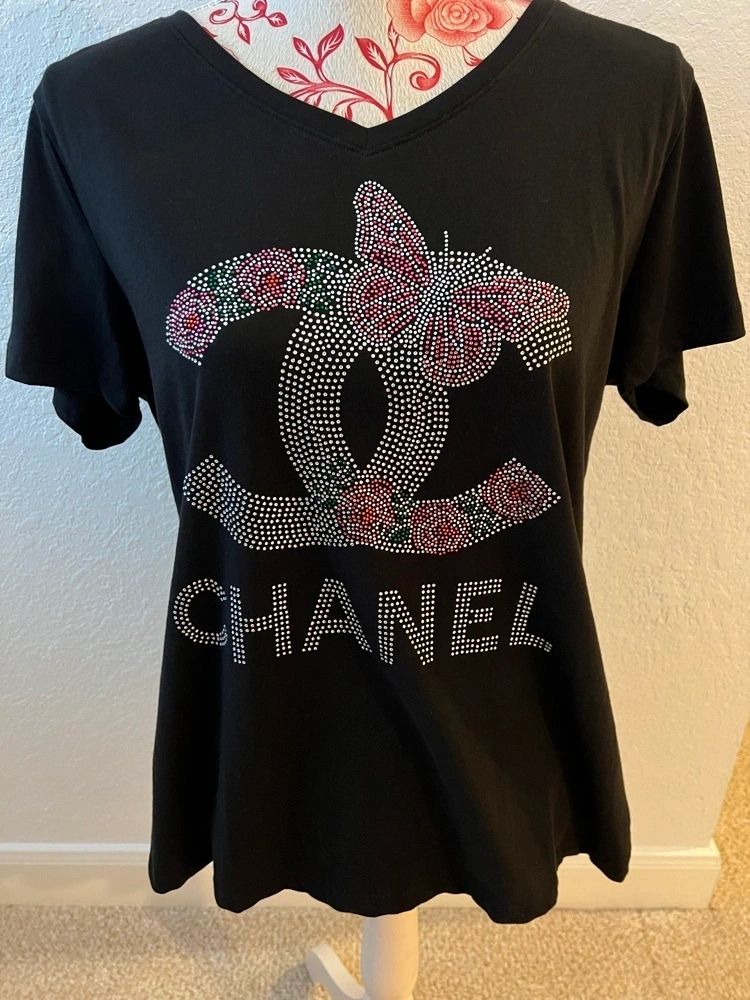 A black women's shirt with a V-neck, featuring a bling-style butterfly and floral design on the front, and the brand logo 'CHANEL' prominently displayed.