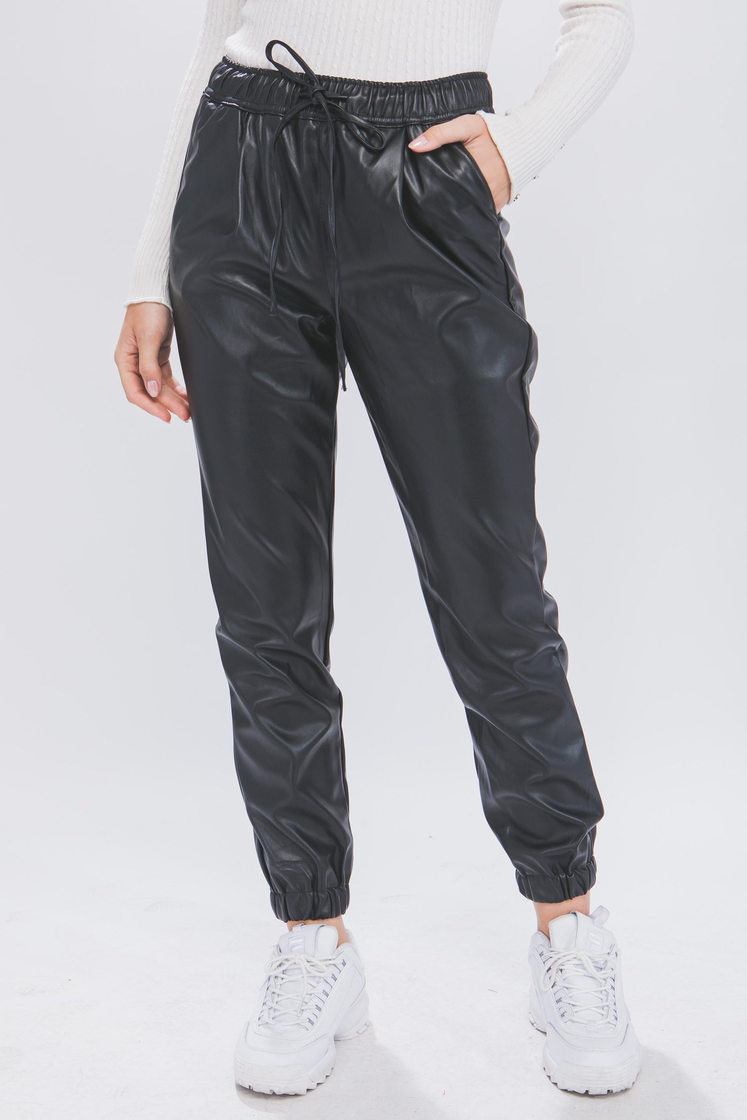 A pair of black faux-leather joggers with an elastic waistband and drawstrings, displayed on a model wearing white sneakers.
