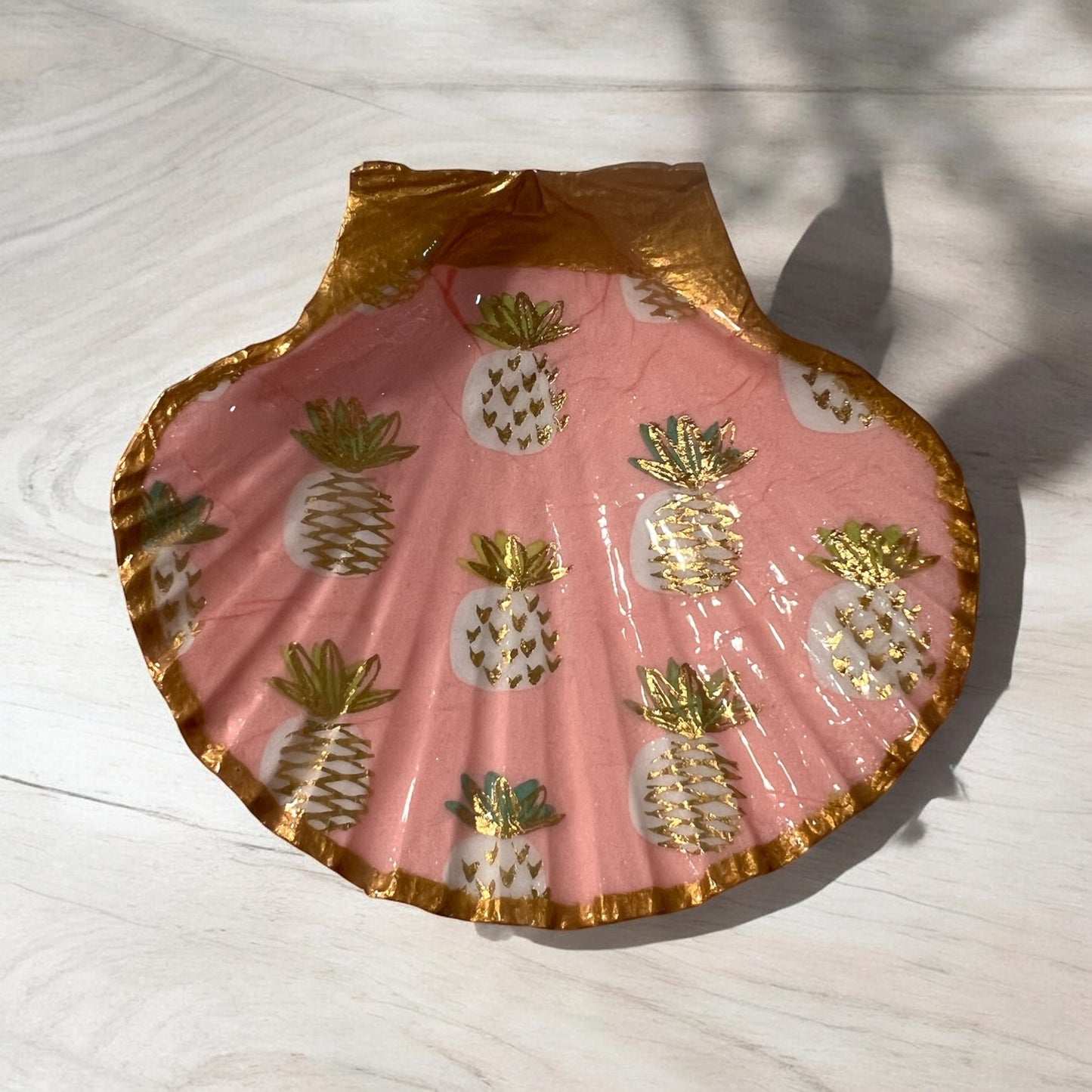 A scallop shell with pink background and gold trim, decorated with pineapple patterns and placed on a wooden surface.