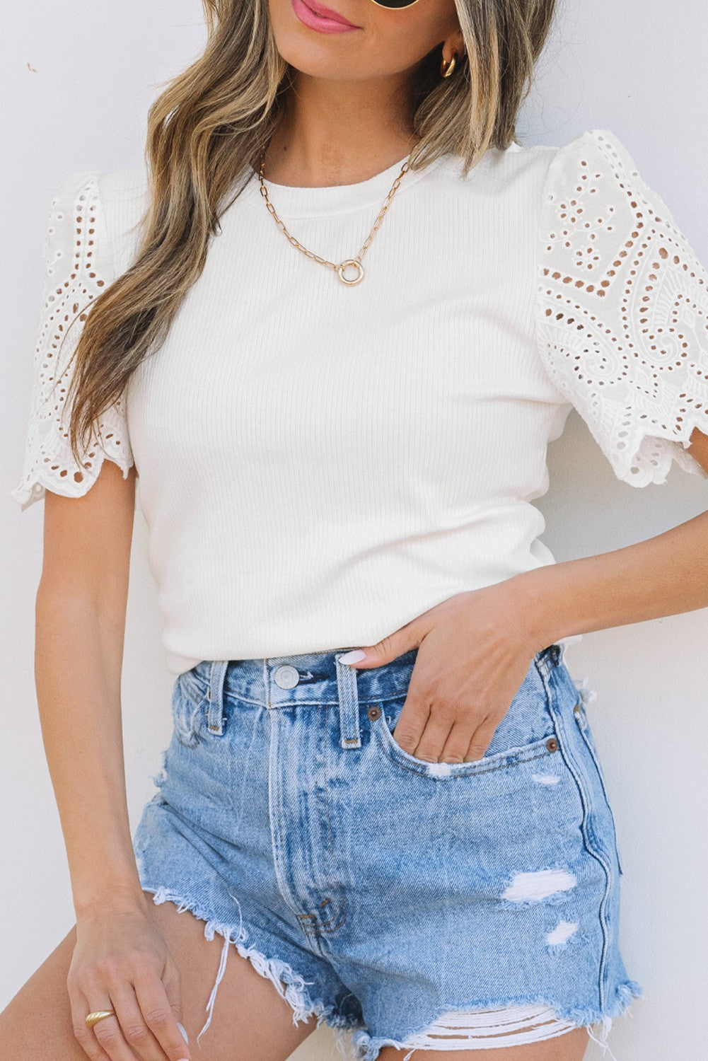 A woman wearing a white eyelet sleeve top with a round neckline and short sleeves, paired with blue denim shorts. She is accessorized with a necklace.